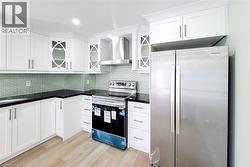 Kitchen featuring stainless steel appliances, wall chimney exhaust hood, decorative backsplash, glass insert cabinets, and white cabinetry - 