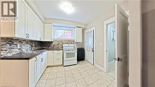 Kitchen with dark countertops, white electric range, white cabinets, radiator heating unit, and decorative backsplash - 25 Spadina Avenue, Hamilton, ON - Indoor Photo Showing Kitchen With Double Sink