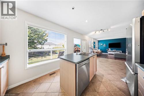 Kitchen featuring dark countertops, an island with sink, appliances with stainless steel finishes, light brown cabinetry, and light tile patterned floors - 132 Blackburn Drive, Brantford, ON - Indoor Photo Showing Kitchen With Double Sink