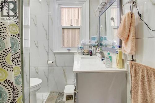 Bathroom featuring a vanity with an integrated sink, a rectangular mirror with integrated lighting, tiled walls, wood-finish flooring, and a single window - 823 Queensdale Avenue E, Hamilton, ON - Indoor Photo Showing Bathroom