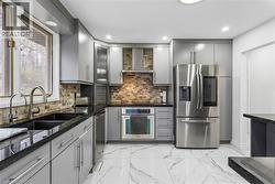 Kitchen featuring gray cabinets, stainless steel appliances, light marble finish flooring, glass fronted cabinets, and dark stone countertops - 