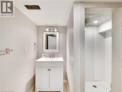 This bathroom features a white vanity with a single sink and a framed mirror, brushed nickel fixtures, and a shower with white tile walls and a mosaic tile floor - 229 Parkdale Avenue N, Hamilton, ON 