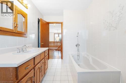 The bathroom features a vanity with a white countertop and a silver faucet, a mirrored medicine cabinet, a bathtub with a shower head, and white tiled walls and flooring - 18 Marcel Place, Hamilton, ON - Indoor Photo Showing Bathroom