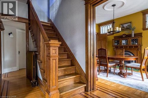 The property offers hardwood flooring with intricate inlay details, a grand wooden staircase with carved newel post, and a room featuring a decorative chandelier and a prominent wooden fireplace mante - 36 Eastbourne Avenue, Hamilton, ON - Indoor Photo Showing Other Room