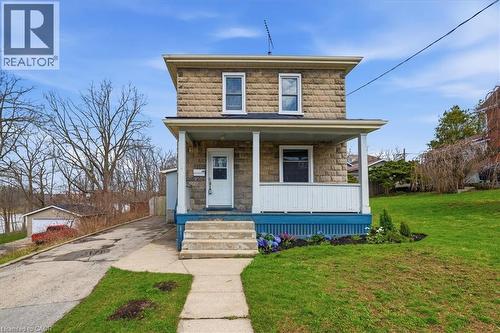 The property features a stone facade, a covered front porch with white railings, and a front yard with a lawn and garden beds - 12 Todd Street, Cambridge, ON - Outdoor With Deck Patio Veranda