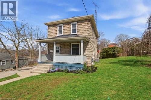 The property features a stone facade, a covered front porch with white columns, and a landscaped front yard with a lawn and flowering plants - 12 Todd Street, Cambridge, ON - Outdoor