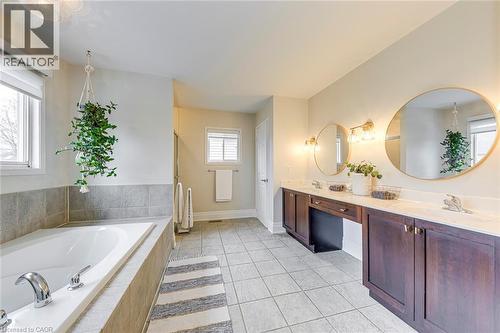 The bathroom features a built-in bathtub with tiled surround, a double vanity with dark wood cabinetry and white countertops, and circular mirrors with wall-mounted light fixtures - 4267 Sarazen Drive, Burlington, ON - Indoor Photo Showing Bathroom
