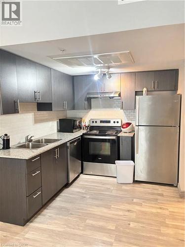 The kitchen features dark wood cabinetry, stainless steel appliances, light-toned countertops, and a white subway tile backsplash - 275 Larch Street Unit# 108, Waterloo, ON - Indoor Photo Showing Kitchen With Double Sink