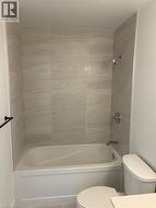 The bathroom features a bathtub with a shower, surrounded by light-colored tiled walls, and a toilet - 