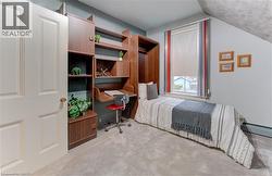 This room features a built-in wooden desk and shelving unit, a window with a roller blind, and light gray carpet flooring - 