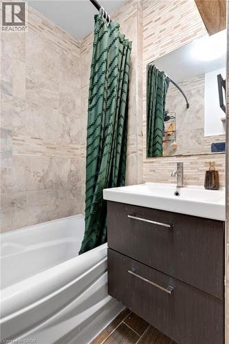 Bathroom featuring a white bathtub, full-height wall tiling, a rectangular mirror, a wall-mounted vanity with two drawers, and wood-finish flooring - 34 Christopher Drive, Cambridge, ON - Indoor Photo Showing Bathroom