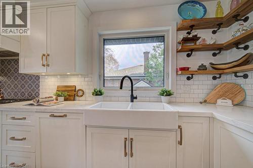 Kitchen featuring a double basin farmhouse sink with a matte black faucet, light-toned countertops, white shaker cabinetry with gold-tone hardware, white subway tile backsplash, and wood-finish open s - 1486 Augustine Drive, Burlington, ON - Indoor Photo Showing Kitchen With Double Sink