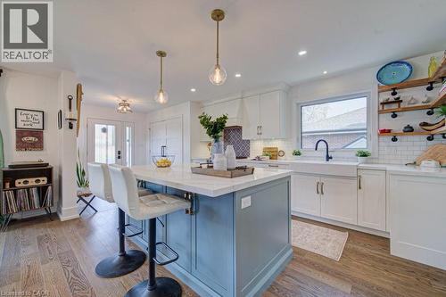 Kitchen featuring a central island with a light blue base and a light-toned countertop, white cabinetry with dark hardware, a white farmhouse sink with a black gooseneck faucet, and wood-finish floori - 1486 Augustine Drive, Burlington, ON - Indoor Photo Showing Kitchen With Upgraded Kitchen