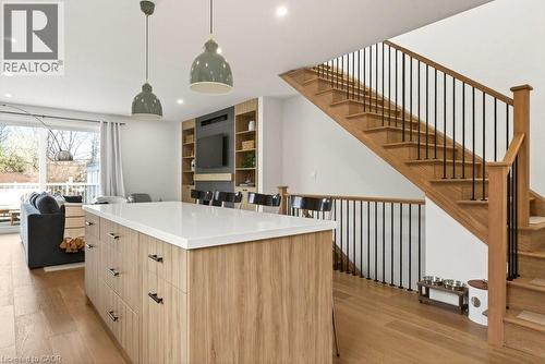 Open-concept living space featuring wood-finish flooring, a contemporary kitchen island with a light-toned countertop, a built-in media center with shelving, and a prominent staircase with wood treads - 128 Canada Street, Hamilton, ON - Indoor
