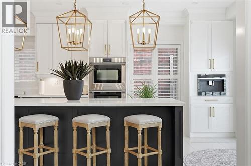 Kitchen featuring a dark-toned island with a light-toned countertop, white cabinetry with gold-toned hardware, built-in stainless steel oven, and two geometric gold-toned pendant lights - 47 Winners Way, Binbrook, ON - Indoor