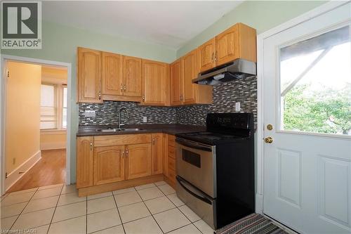 Kitchen featuring light wood cabinetry, a mosaic tile backsplash, dark countertops, a stainless steel range with an overhead vent, and neutral tile flooring - 205 Duke Street E, Kitchener, ON - Indoor Photo Showing Kitchen With Double Sink