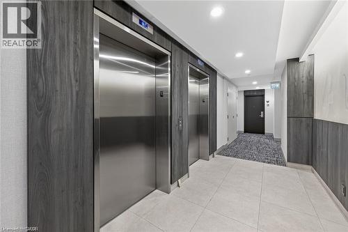 Hallway featuring two stainless steel elevators framed by wood-finish paneling, light-colored floor tiles, recessed lighting, and a textured gray carpet leading to a dark entry door - 85 Duke Street W Unit# 507, Kitchener, ON - Indoor Photo Showing Other Room