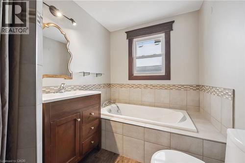 Soaking tub with tiled surround, wood-finish vanity with countertop sink, decorative wall mirror, and window with dark wood trim - 838 Moore Street, Cambridge, ON - Indoor Photo Showing Bathroom