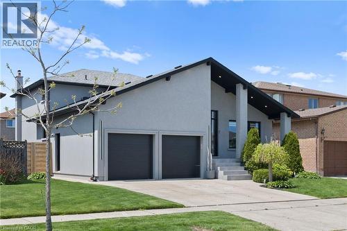 Modern stucco exterior with a prominent A-frame entry, featuring a two-car garage with dark doors, a concrete driveway, and landscaped front yard with mature shrubbery and a young tree - 17 Clearview Drive, Hamilton, ON - Outdoor