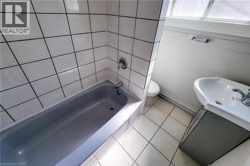 Bathroom featuring a bathtub with white tiled surround, chrome fixtures, a pedestal sink with chrome faucet, a toilet, and a window for natural illumination - 254 Emerald Street N Unit# Upper, Hamilton, ON - Indoor Photo Showing Bathroom