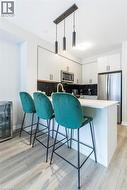 Modern kitchen featuring an island with white countertop, contemporary pendant lighting, light wood-finish flooring, white cabinetry, and stainless steel appliances - 