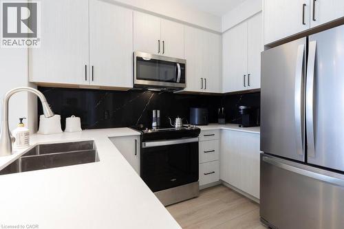 Contemporary kitchen featuring white cabinetry with dark hardware, a stainless steel appliance suite, black marble-patterned backsplash, and light wood-finish flooring - 120 Springvalley Crescent Unit# 208, Hamilton, ON - Indoor Photo Showing Kitchen With Stainless Steel Kitchen With Double Sink