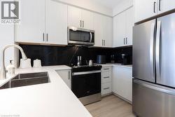 Contemporary kitchen featuring white cabinetry with dark hardware, a stainless steel appliance suite, black marble-patterned backsplash, and light wood-finish flooring - 