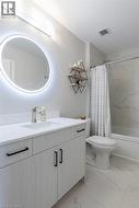 Modern bathroom featuring a single vanity with light wood-finish cabinetry and black hardware, a pristine white countertop with an integrated sink, and an illuminated circular mirror - 