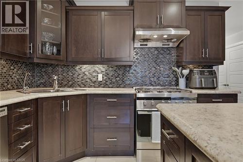 Dark wood-finish cabinetry featuring glass-front display cabinets, stainless steel appliances, a mosaic tile backsplash, and light-toned countertops - 7 Gibson Place Unit# 26, St. Catharines, ON - Indoor Photo Showing Kitchen With Upgraded Kitchen