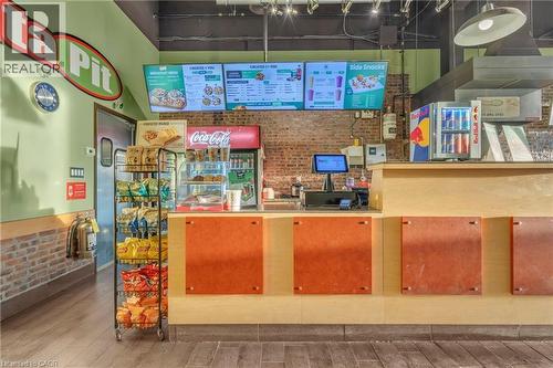 Sales counter area featuring a light wood-finish counter with orange panel accents, digital menu boards, exposed brick wall, and overhead track lighting - 589 Fairway Road S, Kitchener, ON 