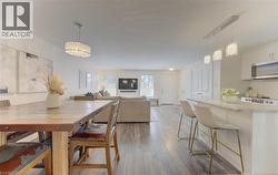 Open-concept living space featuring wood-finish flooring, a kitchen island with seating, pendant lighting, white cabinetry, and stainless steel appliances - 