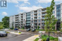 Contemporary multi-story building featuring a gray and white facade, expansive windows, individual glass-paneled balconies, a prominent glass-enclosed entrance, and an asphalt parking area with marked - 