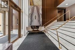Lobby entrance featuring automatic glass doors, wood-finish paneling with brass accents, a large-format abstract art piece, and a grand staircase with marble-look treads and brass railings - 
