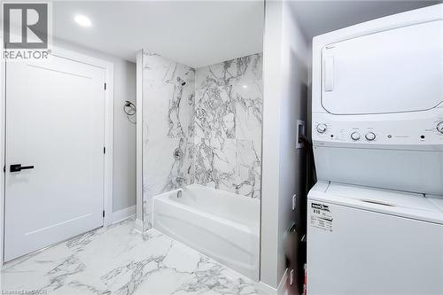 Bathroom featuring a white bathtub with a marble-finish surround, coordinating marble-finish floor tiles, a modern white door with black hardware, and an integrated stacked laundry unit - 103 East 45Th Street, Hamilton, ON - Indoor Photo Showing Laundry Room
