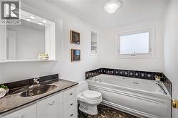 Bathroom featuring a jetted tub, a single vanity with a dark granite countertop, a wall-mounted mirror with integrated lighting, a window, and built-in shelving - 