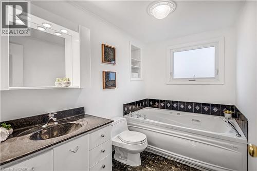 Bathroom featuring a jetted tub, a single vanity with a dark granite countertop, a wall-mounted mirror with integrated lighting, a window, and built-in shelving - 85 Ranchdale Drive, Hamilton, ON 