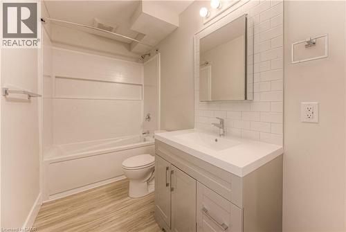 Bathroom featuring a white subway tile backsplash, a single vanity with a white countertop and gray cabinetry, a white bathtub with a shower kit, and wood-finish flooring - 40 Daniel Avenue Unit# 2, Kitchener, ON - Indoor Photo Showing Bathroom