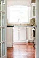 Kitchen area featuring wood-finish flooring, white cabinetry with gold-tone hardware, a large window with architrave detailing, a stainless steel faucet, and dark countertops - 