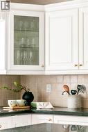 White upper cabinetry with gold-finish hardware, glass-front display cabinet, dark stone countertops, and a light-toned square tile backsplash - 