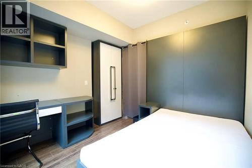 Room featuring wood-finish flooring, a built-in desk with open shelving, a wall-mounted shelving unit, and a built-in wardrobe with a white and gray finish - 330 Phillip Street Unit# S605, Waterloo, ON - Indoor Photo Showing Bedroom