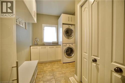 Dedicated laundry area featuring stacked appliances, a utility sink, a window with blinds, upper and lower cabinetry, and tiled flooring - 711 Cedar Bend Drive, Waterloo, ON - Indoor Photo Showing Laundry Room