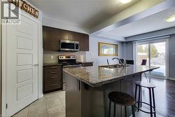 Kitchen featuring an island with stone countertop and undermount sink, stainless steel appliances, dark wood cabinetry, tile backsplash, and sliding glass door access to the exterior - 