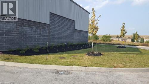 Exterior featuring a gray block wall base, light gray corrugated metal siding, a landscaped planting bed with dark mulch, several young trees, and a concrete curb bordering an asphalt surface - 44 Ditton Drive Unit# 3, Hamilton, ON 
