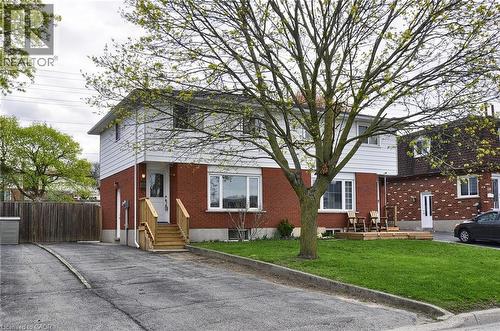 Two-story residence featuring a brick and white siding exterior, a front entry with wooden steps and railing, a front yard with a mature tree and green lawn, a wooden deck, and an asphalt driveway - 114 Bakersfield Drive, Cambridge, ON - Outdoor