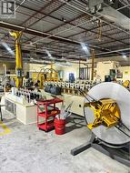 Industrial workspace featuring a metal roll forming line, exposed structural ceiling, integrated overhead lighting, concrete flooring, and yellow safety stanchions - 
