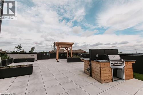 Spacious rooftop patio featuring light-toned large format pavers, a built-in outdoor grill station with wood-finish cabinetry, a wooden pergola, and dark planters with greenery - 251 Northfield Drive E Unit# 406, Waterloo, ON - Outdoor
