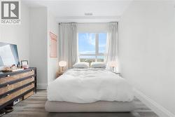 Crisp white walls frame a bright bedroom featuring wood-finish flooring, a large window with white drapery, and recessed lighting - 