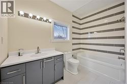 Bathroom featuring a white bathtub with a horizontally striped tile surround, a built-in toilet, a window, a vanity with gray cabinetry and a white countertop, and a six-bulb light fixture - 