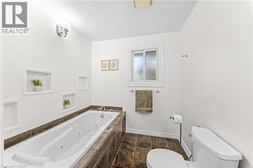 Soaking tub with integrated jets, surrounded by dark tile with a matching floor, featuring wall-mounted recessed shelves, a frosted window, and a white toilet - 183 Harvard Road, Waterloo, ON - Indoor Photo Showing Bathroom