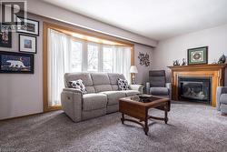 Living area featuring a prominent wood fireplace with a detailed mantel, a bay window with a wood-finish frame, light-toned wall paint, and gray carpet flooring - 